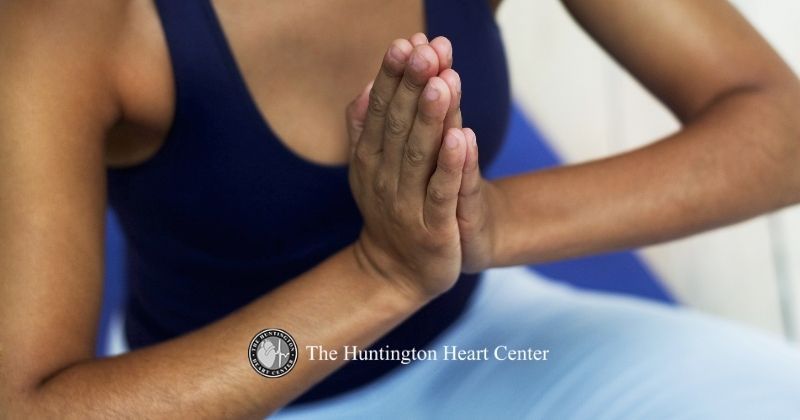 Woman in blue meditates on mat for cardiovascular health and heart benefits with hands at heart center. Huntington Heart Center logo at bottom center.