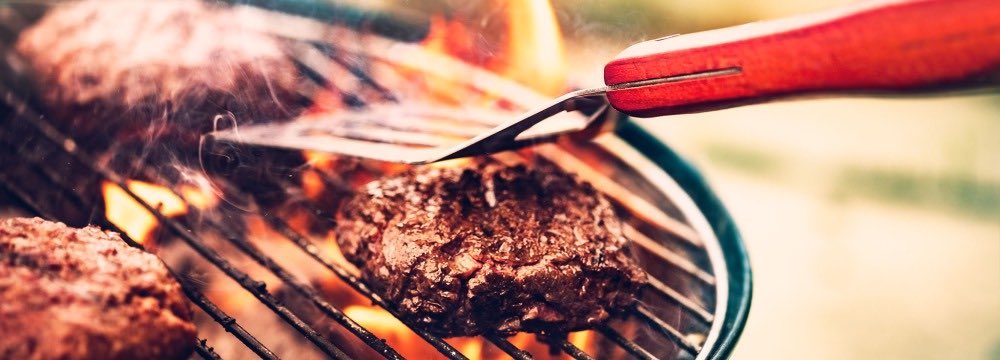 Burger being cooked on a grill with flames and spatula